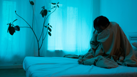 A depressed person alone in their room sitting on their bed with a blanket over their head. 