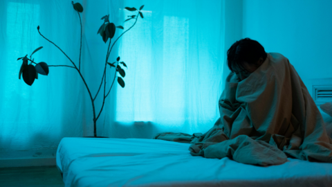 A depressed person alone in their room sitting on their bed with a blanket over their head. 
