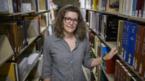 A librarian stands in the aisle between library shelves, with one arm resting on a book