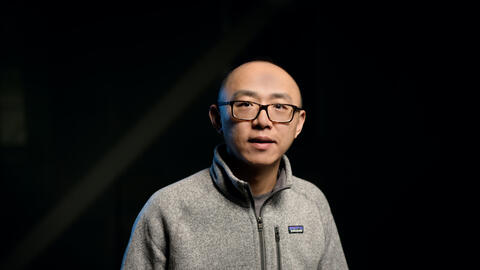 Jiayu Zhou wears a sweater and a black pair of glasses and looks at the camera. 