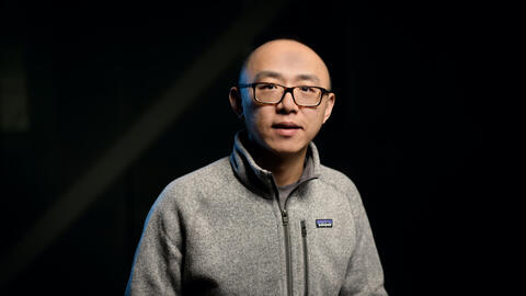 Jiayu Zhou wears a sweater and a black pair of glasses and looks at the camera. 