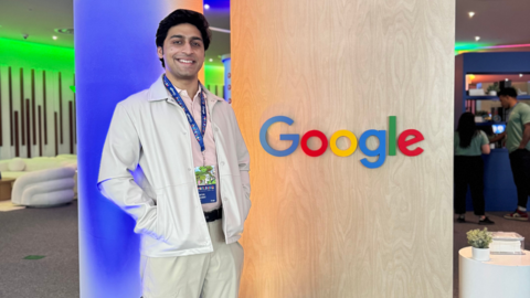 An individual smiles proudly wearing a lanyard and standing in front of a light wood wall adorned with the Google logo