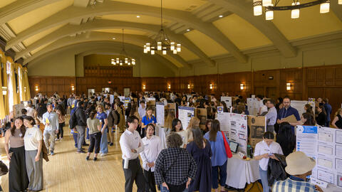 A view of a crowd gathered in a well-lit ballroom with tall ceilings, filled with tables holding posters 