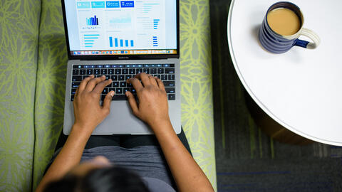 A view from above as a person works on a laptop with graphs visible on the screen. A coffee mug rests on a white table beside them