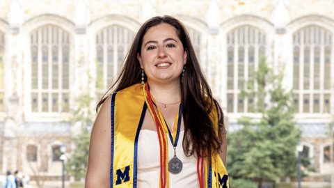 A student wearing a maize stole, red cords, and a medal poses in front of an academic building with tall windows