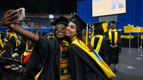 A close-up of two graduates in caps and gowns taking a selfie from the floor of Crisler Center, with rows of graduates and the blue-curtained stage visible behind them