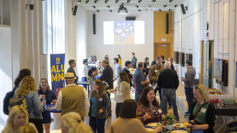 Many people gather in a bright event space with round tables. A banner reads "Alumni," and a slideshow of photos is projected on the back wall