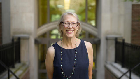 A headshot of Elizabeth Yakel posing in front of the glass entrance to a building, smiling in a navy linen blouse 