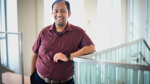 Venkatesh Potluri, a man with short dark hair, wearing a maroon button-up shirt and dark pants, stands indoors leaning casually against a glass railing. He is smiling and has one hand in his pocket, with the other resting on the railing. Soft natural light filters in through large windows behind him.