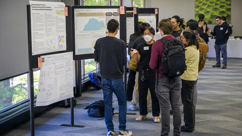 Alt text:  A group of graduate students stand in a bright indoor space, gathered around research posters displayed on black presentation boards.