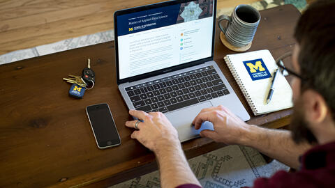 A close-up of a person typing on a laptop with a  University of Michigan notebook, coffee cup, phone and keys surrounding it.