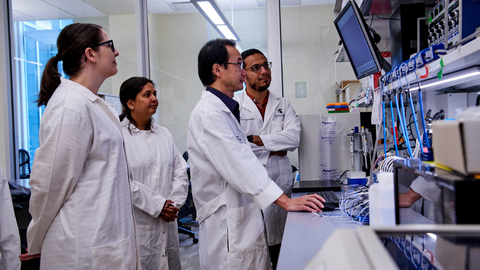Four researchers in white lab coats stand together in a laboratory, looking at a computer screen and equipment on a bench.