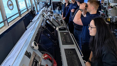 Students and U.S. Coast Guard personnel stand inside a vessel’s bridge, surrounded by navigation equipment and control panels, as one person looks through binoculars and others observe the water through large windows.