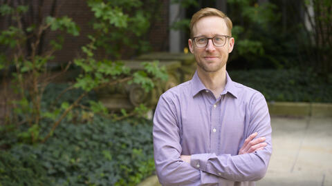 Card wearing glasses and a light purple button-down shirt standing outdoors with his arms crossed, looking at the camera.