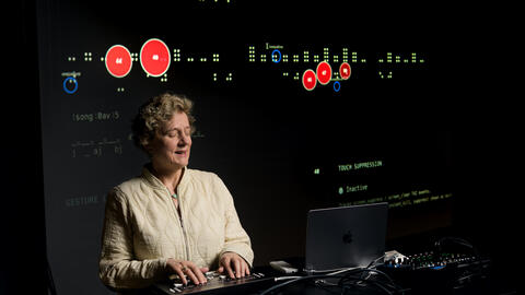 Sile is brightly illuminated amid a large dark studio space, standing at a podium and interacting with a refreshable braille display. A large projection screen behind her shows green braille dots as a representation of the screen state, and red dots for where the screen is being touched. The green braille dots show a file name, blocky audio waveform, playhead position and timecodes in seconds.