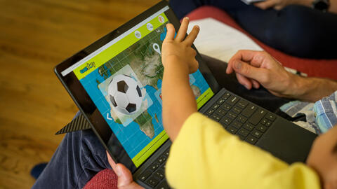 A child reaches to press a screen that displays a map and an image of a soccer ball. An adult holds the laptop.