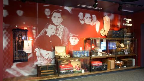 Physical exhibit at the Arab American National Museum titled “Coming to America” features a display of luggage and personal items, with a large monochrome photo of a family on the back wall.