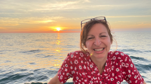 Barbara Ericson smiles as the sun sets over a watery horizon at her back. 