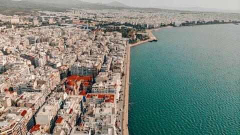 An aerial view of the Port of Thessaloniki depicting clear blue waters on the right and clustered buildings, some modern and some historic with red roofs, on the left.