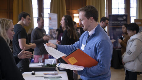 A student holding an orange folder hands a resume to a recruiter standing on the other side of a table.