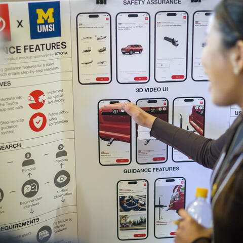 A student gestures toward a project poster with mockups of mobile designs.