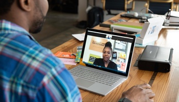 A person sits at a table with a laptop that is open to a virtual meeting with another person on their screen.