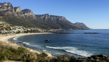 Landscape photo of Cape Town, South Africa coastline. The image captures people on the shore and in the water, and buildings below jagged cliff sides. 