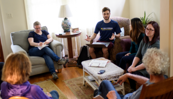A group of six people sit on armchairs and wooden chairs around a coffee table in a homey environment. One person takes notes in a notebook, and others listen to a person wearing a University of Michigan School of Information T-shirt who gestures openly while balancing a laptop on their lap. 