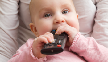 Baby wearing a long-sleeve pink shirt and holding a remote to their mouth. 