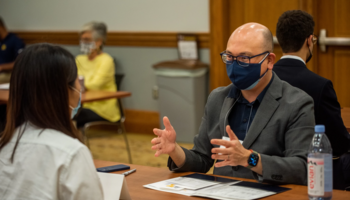 Alumnus Rodger Devine sits across the desk from a current UMSI student. He is masked but speaking animatedly. 