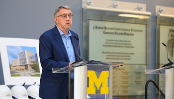 Donor Larry Leinweber speaks at a podium adorned with a Block M. Behind him are commemorative hardhats and a rendering of the Leinweber Computer Science and Information building. He is wearing glasses and a blue blazer. 