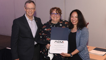 Tiffany Veinot is flanked by the past and present presidents of ACMI. They are smiling and holding an award. 