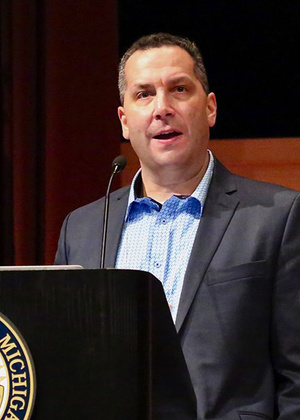 A headshot of Sol Bermann at a podium with the University of Michigan seal on it.