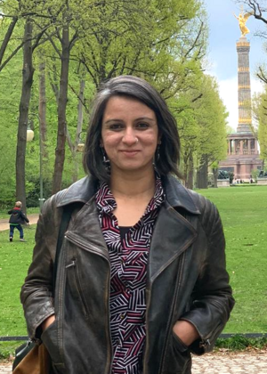 A portrait of Anubha Singh standing in a park. There are trees and a monument behind her.