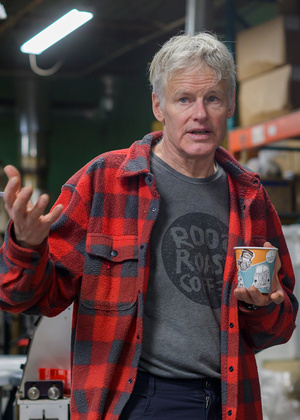 John Roos stands in the roastery, holding a RoosRoast coffee cup and gesturing while speaking