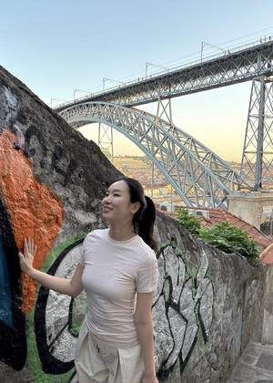 Jiyeon Seo standing on outdoor stairs with her hand on a graffitied wall. There are electric towers and a city in the background.