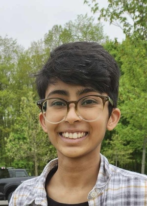 A headshot of Manali smiling outdoors with green trees in the background. They are brown with short black hair, wearing glasses and a grey and white plaid button-down shirt. 