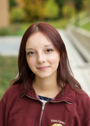 Veronica Pimenova smiling in a red jacket standing outside of the Leinweber Building right before it starts to rain.