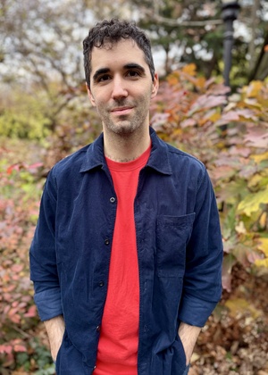 Ben Green wearing a red t-shirt with a blue overshirt, standing in front of fall foliage. 