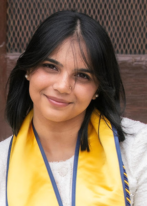 A headshot from Shalini smiling into the camera. She is wearing a white textured blazer and a University of Michigan yellow and navy blue graduation sash. 