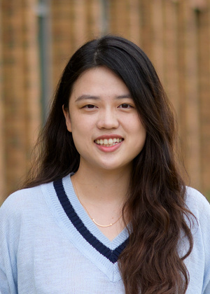 Xin Ye smiles at the camera in an outdoor portrait, with a softly blurred wooden background. She has long dark hair and is wearing a light blue V-neck sweater with navy trim and a delicate gold necklace.