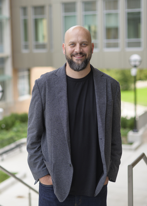 A professional portrait of James Rampton standing outdoors on the University of Michigan campus. He is smiling and wearing a gray blazer, black shirt, and jeans, with modern university buildings, landscaped walkways, and stairways softly blurred in the background.