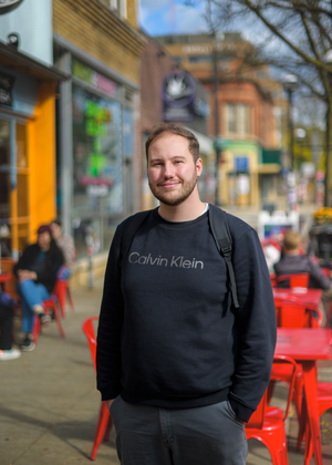 Joey Quick stands on the sidewalk in front of RoosRoast coffee in downtown Ann Arbor, smiling at the camera