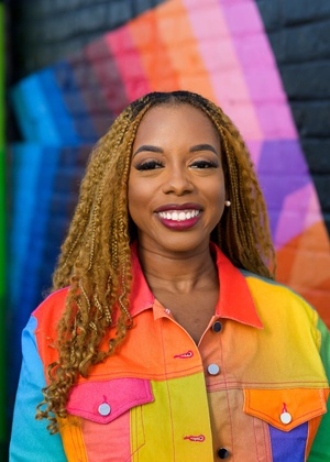 Megan poses in front of a rainbow and prismatic mural in an alleyway while wearing a vibrant dress and jacket of a patchwork of shapes and colors.