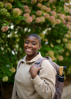 Tyra Briscoe smiles in front of a flowering bush. She wears a backpack with a duck umbrella in the pocket.
