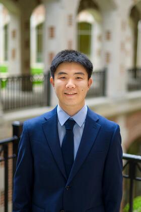 Jiaxin Pei wearing a blue suit and tie, standing outside and smiling at the camera. 