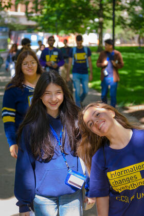In the foreground, two students smile for the camera while wearing U-M blue. The first wears a t-shirt that says "Information changes everything." Other CCSI participants are visible walking behind them.