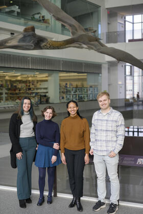 Madison Haack, Julia Cave Arbanas, Cameron Robertson and Austin Book pose on the second level of a museum with a life-size model of a prehistoric pterosaur visible behind them
