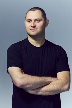 A headshot of Mark Gurman posing with arms crossed against a slate gray background