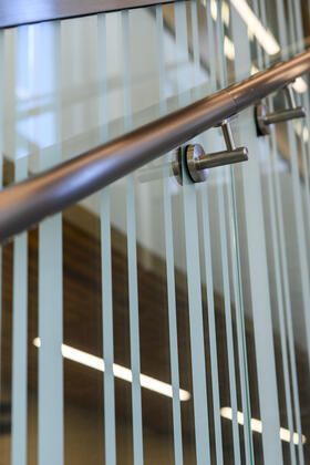 A close-up of a glass railing with a silver rail. The railing has a frosted glass barcode motif all along it